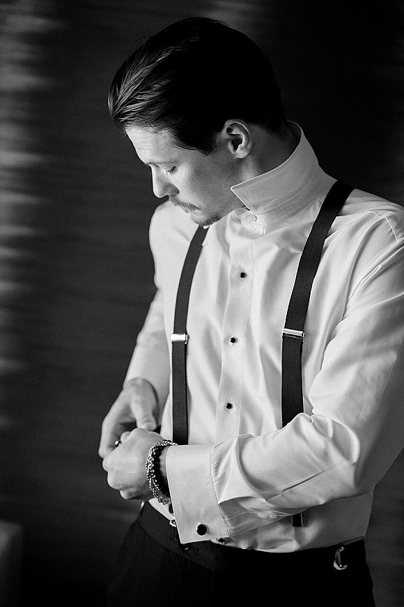 Groom getting ready, putting on cufflinks over a white dress shirt with black suspenders and belt against a dark wall