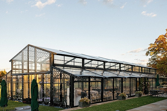 Clear tent reception with a black frame set on a stone patio, with outdoor tables, chairs, and umbrellas on the lawn under clouds