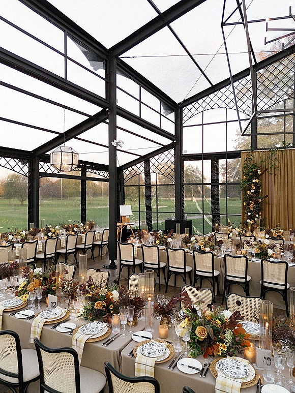 Reception tablescape in a glasshouse wedding reception with long banquet tables, floral garland, taper candles, and dusk views through window walls