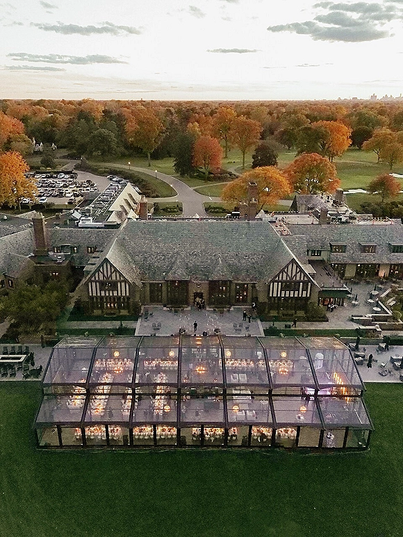 Wedding reception tent with a clear top, string lights and chandeliers over round tables with white linens on an estate lawn