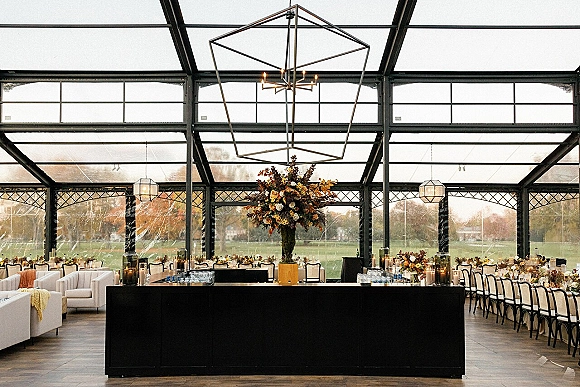 Reception decor with round dining tables and candlelit tall floral centerpieces beside a black bar in a glass wall venue with lawn view