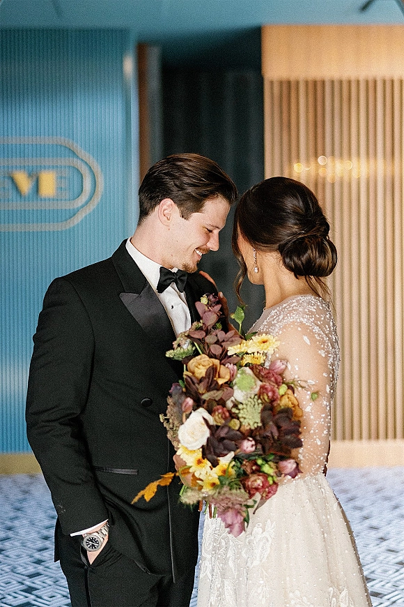 Couple portrait of bride and groom portrait with foreheads touching, her beaded gown and bouquet in a paneled hallway with patterned carpet