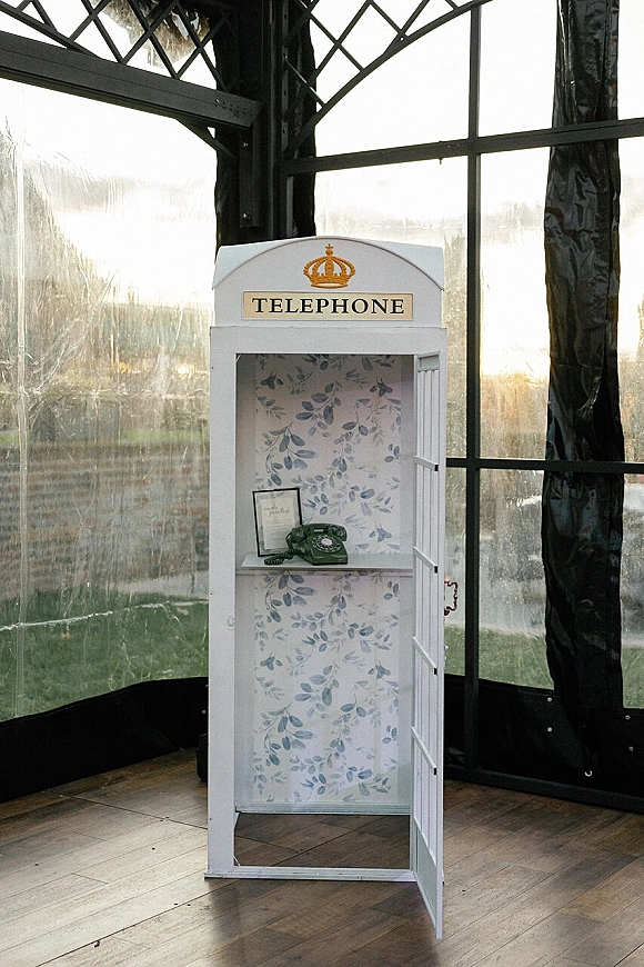 Wedding phone booth audio guestbook station with vintage rotary phone and floral wallpaper, open in a glass greenhouse with greenery beyond