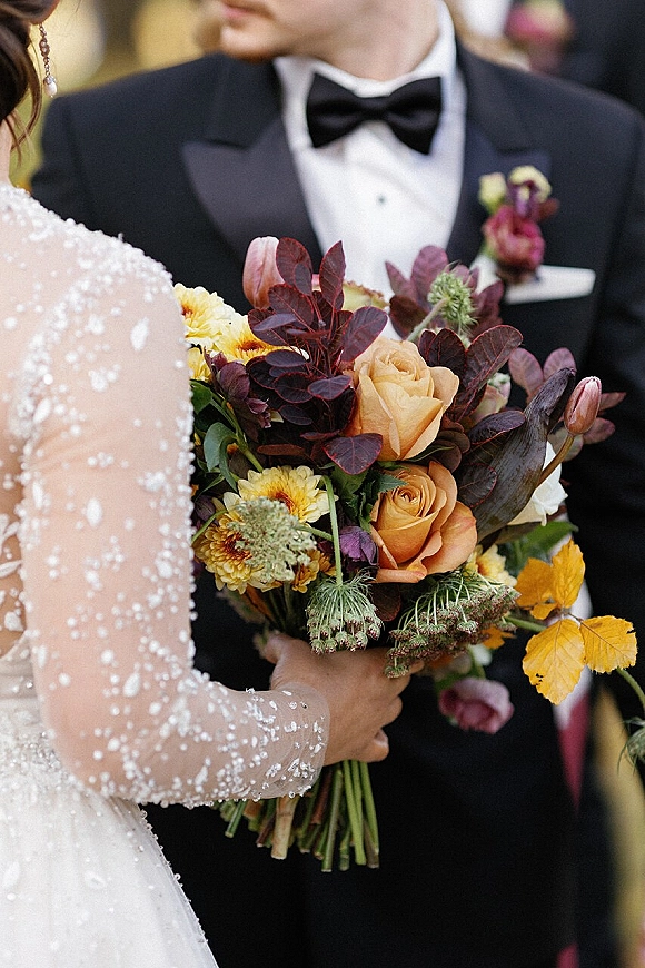 Bridal bouquet with orange roses and fall wedding bouquet tones, held by bride in beaded lace sleeves beside groom in black tuxedo outdoors