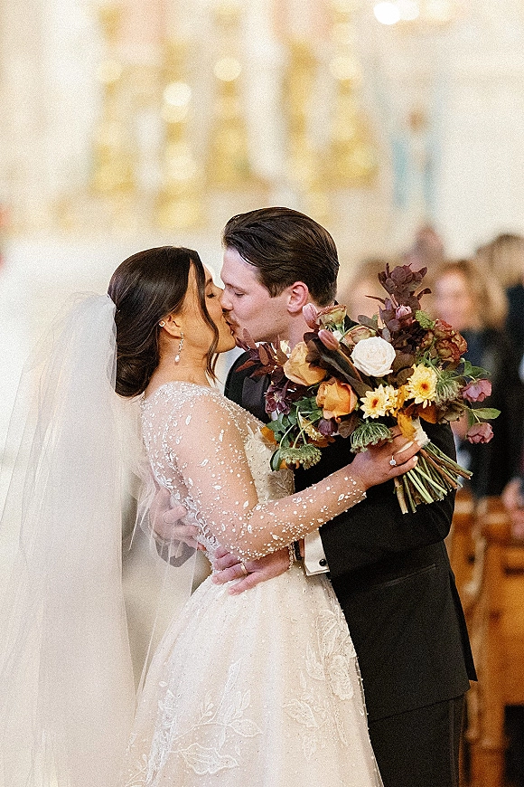 Wedding kiss portrait of bride and groom kissing, bouquet in hand, with bridal veil and warm bokeh lights in an indoor ceremony space