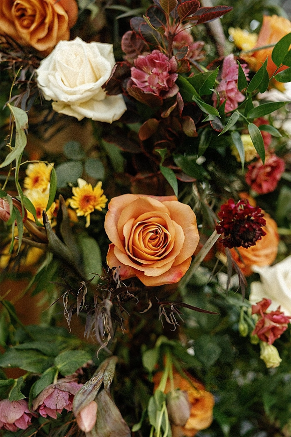 Wedding floral arrangement with rust and orange roses, mixed blooms and greenery, set against a softly blurred floral backdrop