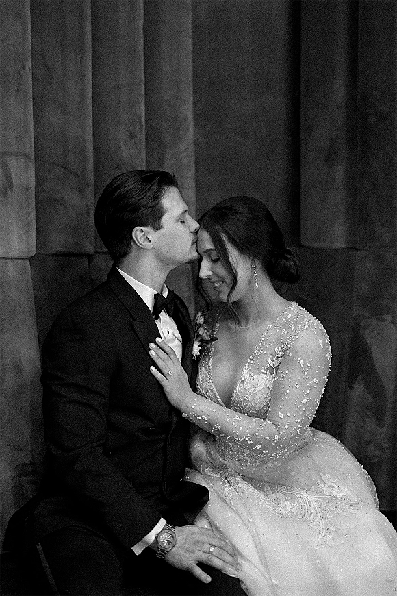 Couple portrait of groom kissing bride’s forehead in a close embrace, her long sleeve lace dress against stone wall and columns backdrop