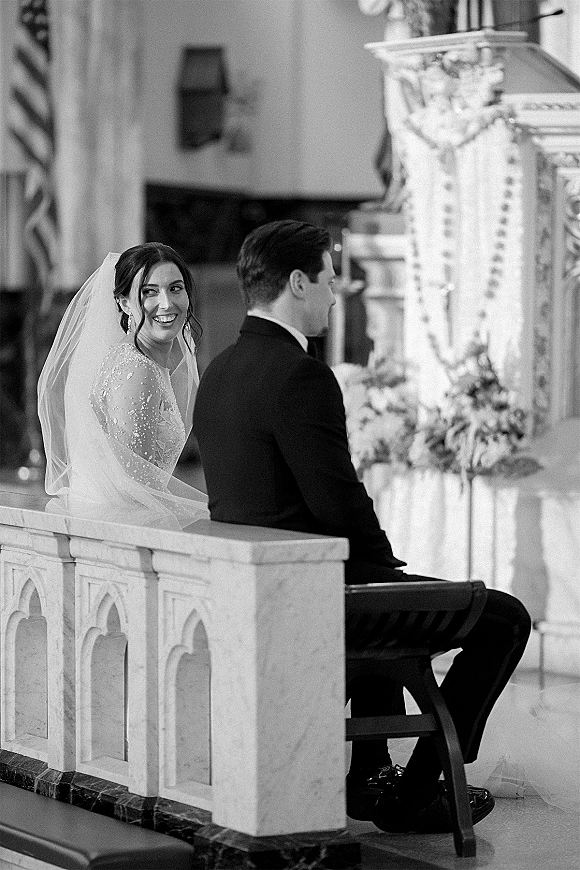 Ceremony moment in a black and white wedding ceremony, bride smiling back at groom with cathedral veil as they sit at the altar in church interior