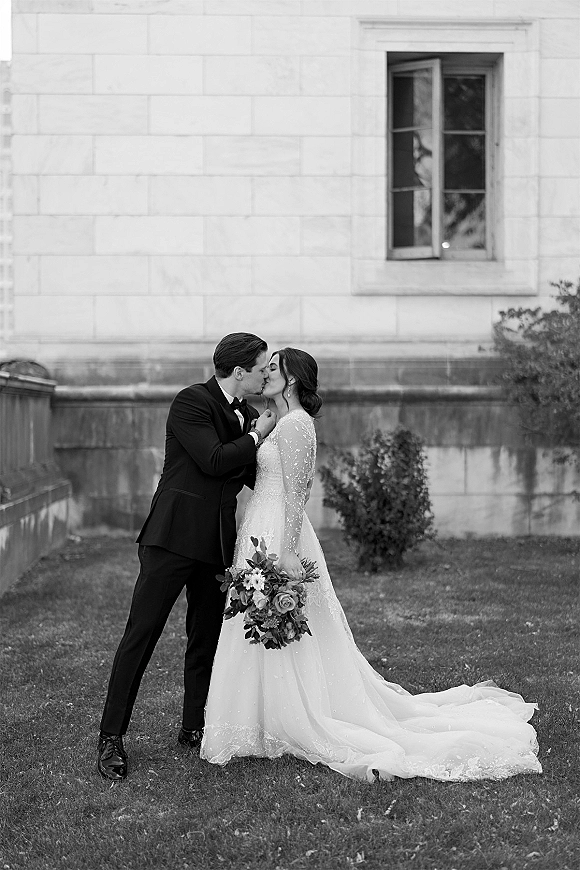 Wedding kiss portrait of bride and groom kissing, her long-sleeve lace dress and bouquet against a stone building wall and lawn backdrop