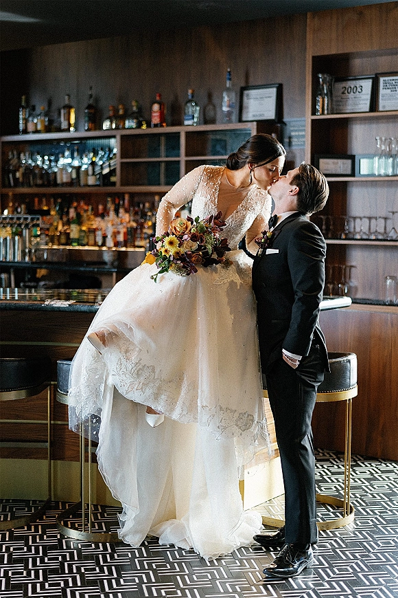 Wedding kiss portrait of bride sitting on a bar as groom leans in, her beaded long-sleeve gown and bouquet against a liquor shelf backdrop