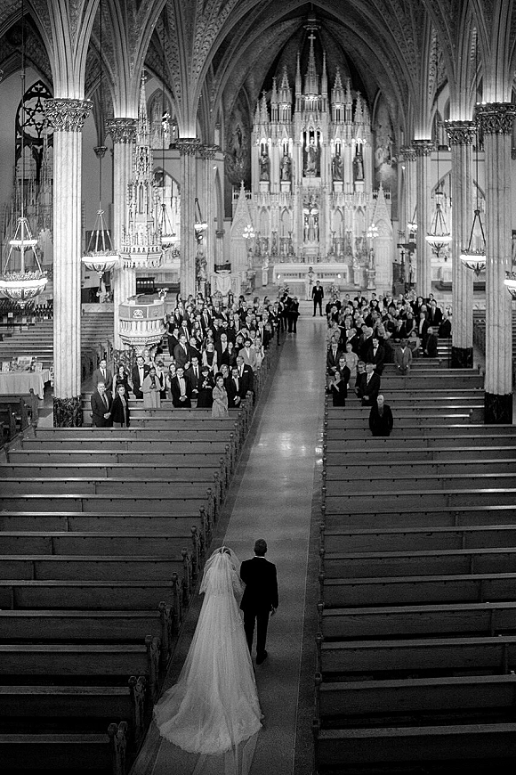 Wedding processional as bride walks down aisle from behind, long veil and train flowing toward candlelit altar in grand cathedral interior