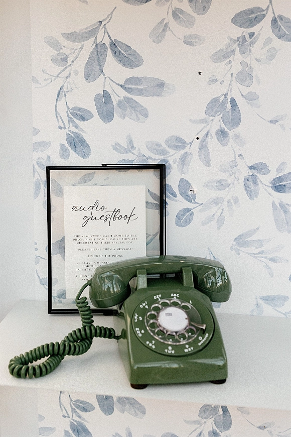 Wedding audio guestbook with an audio guestbook phone, green vintage rotary handset beside a framed sign on a white shelf with leaf wallpaper