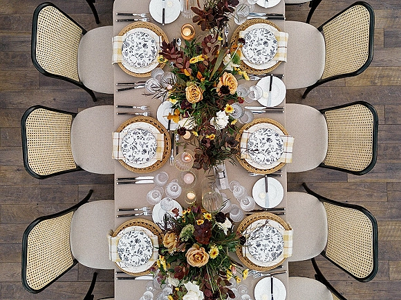 Reception tablescape wedding table setting with roses and greenery garland, rattan chargers, patterned plates, plaid napkins, and votives on wood floor