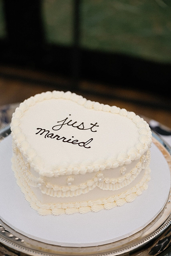 Wedding cake with just married icing in cursive on white frosting, finished with a piped border on a silver stand indoors