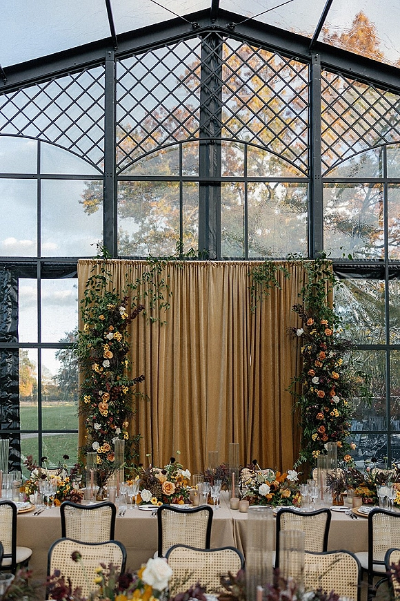 Reception head table with a gold drape backdrop, floral pillars, and greenery garland, set with taper candles and champagne coupes in a glass greenhouse