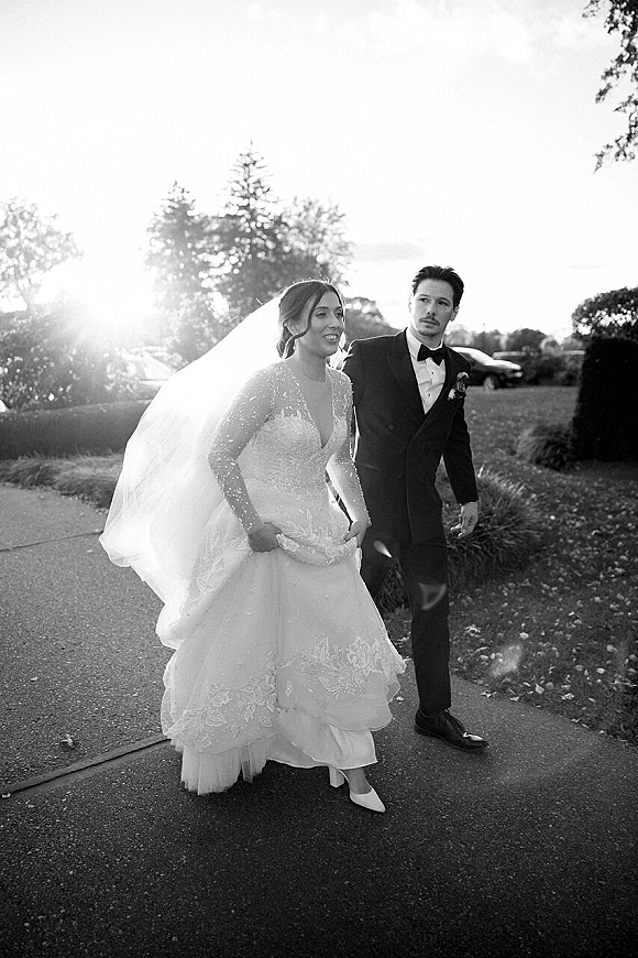Wedding couple portrait of bride and groom walking on a sunlit path, her long veil trailing as he looks at her in a tuxedo