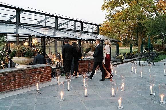 Reception entrance with a candlelit wedding walkway lined with glass cylinder candles and lanterns on a stone patio by a glass greenhouse at dusk