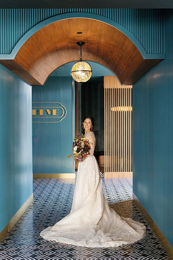 Bridal portrait of a bride holding bouquet in a long sleeve wedding dress, looking over her shoulder beneath an arched blue hallway doorway