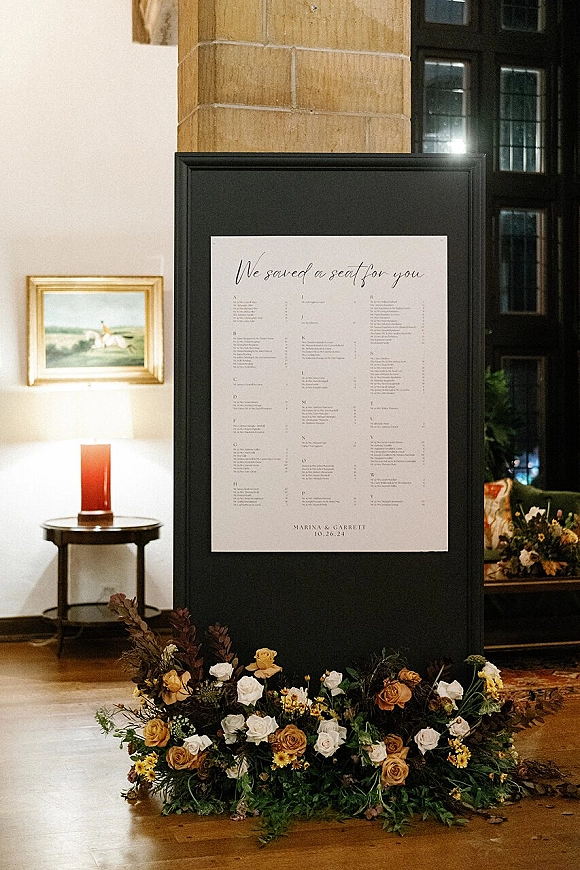 Wedding seating chart in a framed seating chart sign with calligraphy, flanked by a floral arrangement and candle near tall windows indoors