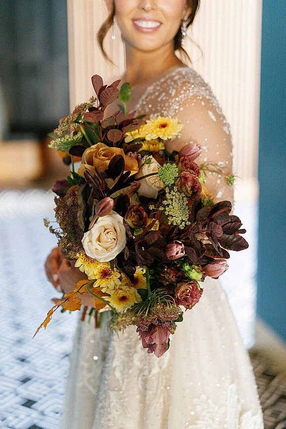 Bridal bouquet with a fall wedding bouquet palette of roses, tulips, and chrysanthemums, held against a beaded dress in window light