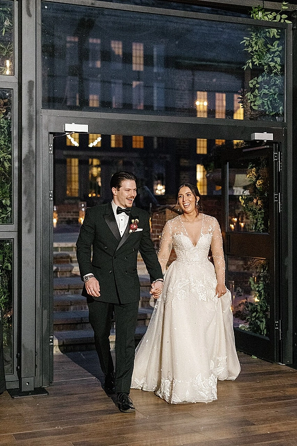 Newlywed couple portrait of bride and groom holding hands, smiling at a reception entrance with greenery garland and candlelit glass doors