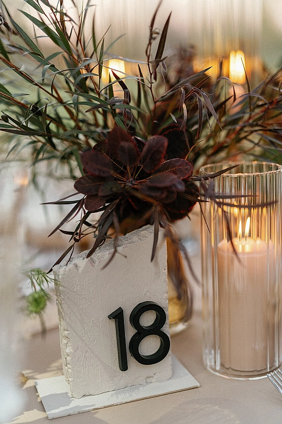 Wedding table number centerpiece with black numbers beside a floral and greenery arrangement and glass candle holder in warm reception light