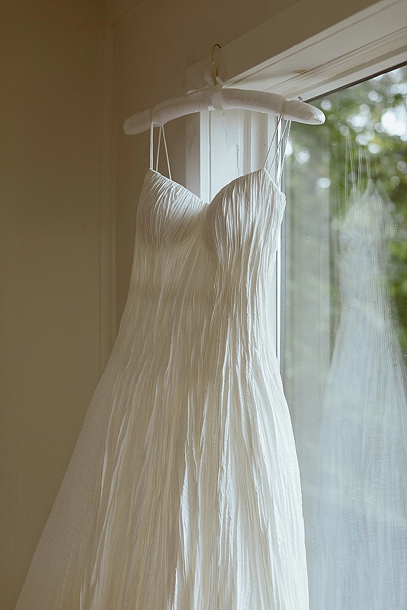Wedding dress hanging in a window on a padded hanger, ribbon bow at the top, with soft daylight and greenery outside