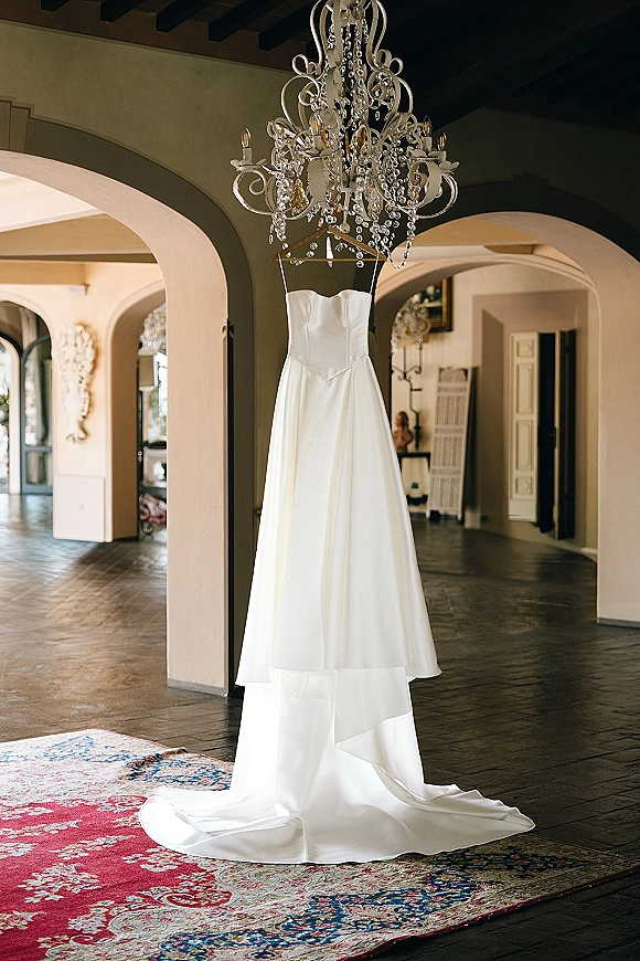 Wedding dress hanging from a chandelier, strapless satin gown with long train in an interior hallway with arched doorways and beams