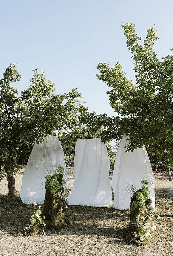 Ceremony backdrop with white draped fabric and moss-covered pedestals topped with green hydrangeas and white florals in an orchard meadow setting