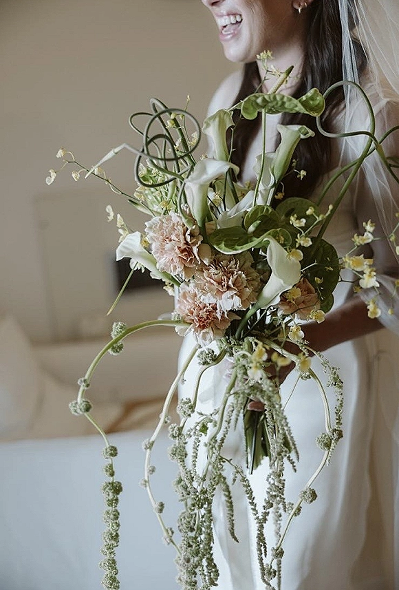 Bridal bouquet with calla lilies and hanging amaranthus, held by a veiled bride in a strapless dress in a neutral room