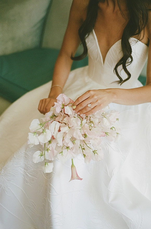 Bridal portrait of a bride holding bouquet in a strapless wedding dress, showing her engagement ring while seated on a green sofa indoors