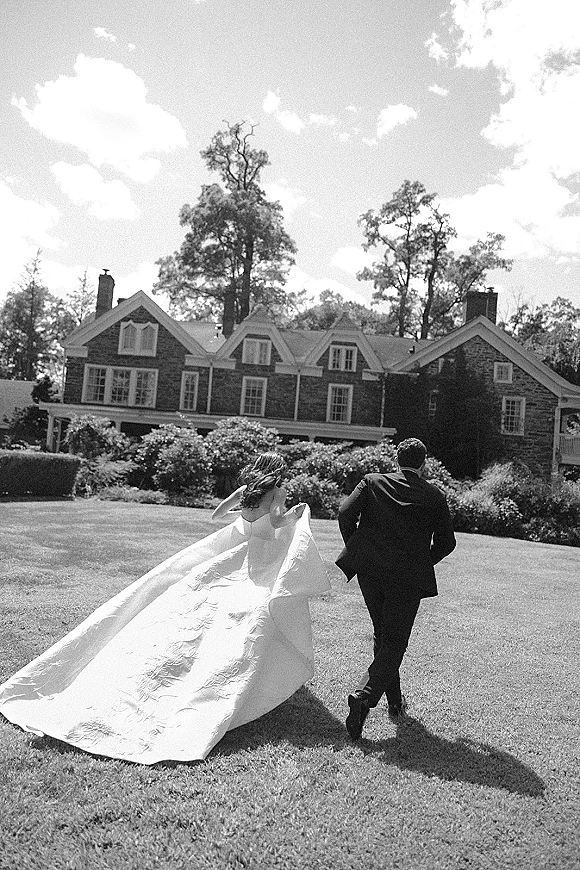 Wedding couple portrait in a black and white wedding photo, bride in strapless gown with long train walking with groom on lawn by stone house