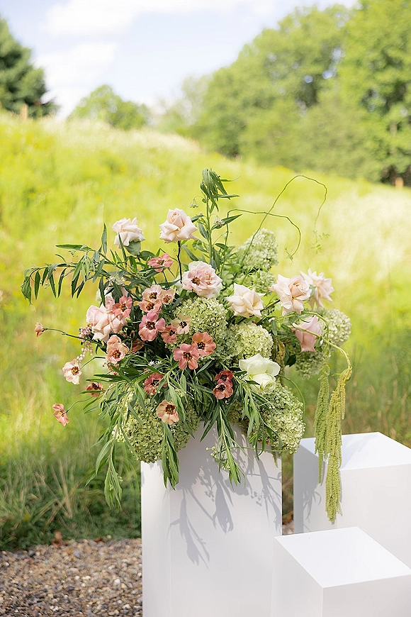 Wedding floral arrangement on a ceremony floral pedestal with blush roses, pink blooms, and green hydrangea beside meadow grass and trees