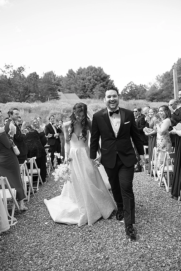 Wedding recessional as bride and groom walking aisle hand in hand, bride in long veil holding bouquet, guests cheering in outdoor field