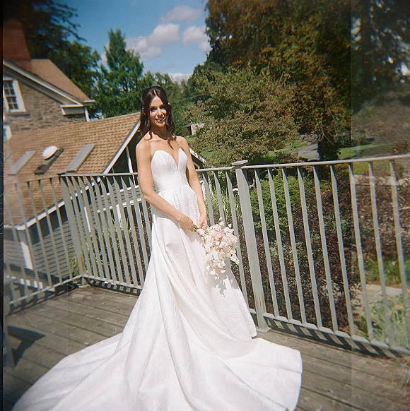 Bridal portrait of a bride in a strapless wedding dress holding a pastel pink bouquet on a wood deck by a stone building under blue sky