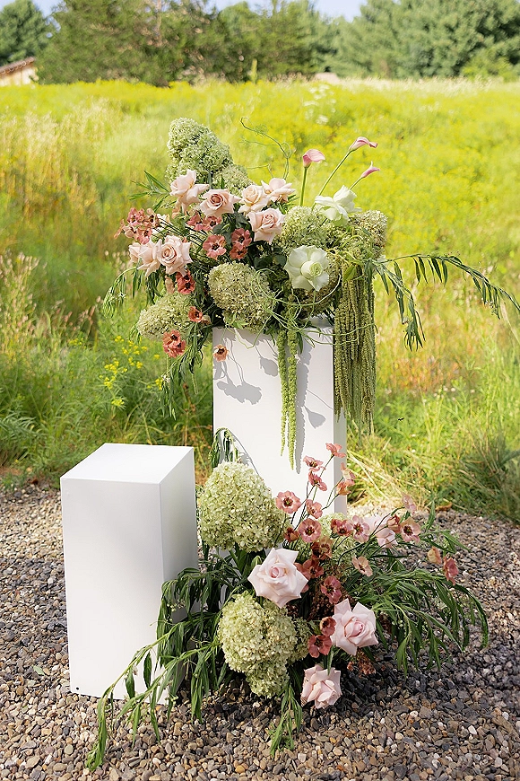 Wedding floral pedestal on white plinths with blush roses, hydrangea and trailing amaranthus in a wildflower field setting