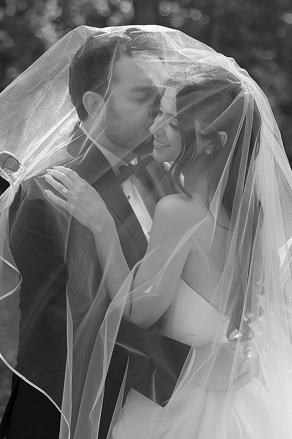 Wedding couple portrait with veil draped over them as groom kisses bride’s forehead, her engagement ring visible amid soft bokeh trees