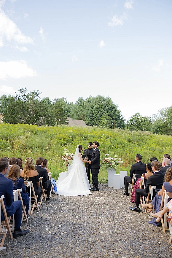 Wedding vows as bride and groom face the officiant beneath a floral arch, bride’s cathedral veil trailing along a gravel aisle in a meadow