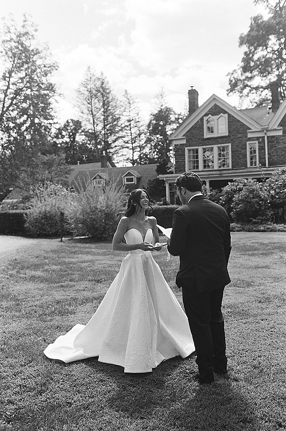 Wedding vows as bride and groom read paper vows facing each other, her strapless dress and train flowing on a lawn by a brick house