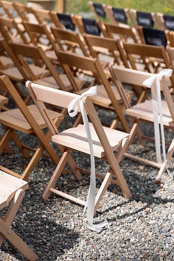 Ceremony seating with outdoor ceremony chairs, wood folding chairs tied with white ribbon bows and reserved signs on gravel beside grass