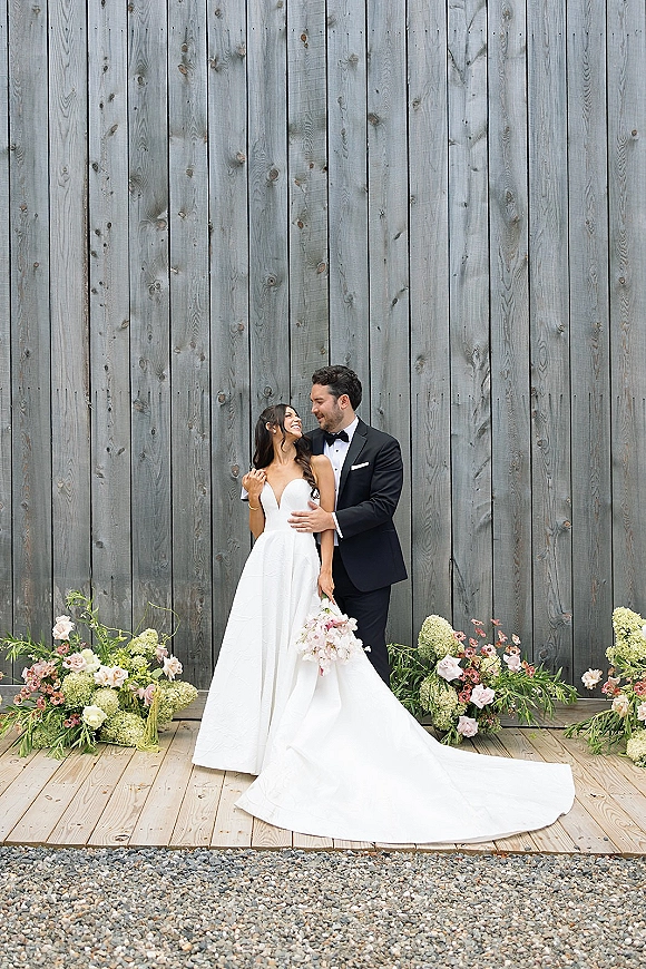 Couple portrait of bride and groom embrace, groom behind bride in strapless gown with long train beside pastel bouquet on weathered wood wall