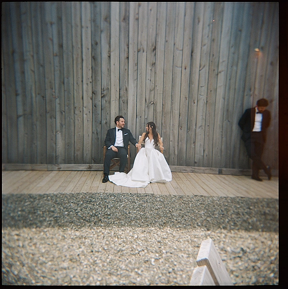 Couple portrait of bride and groom seated holding hands, groom looking at bride, veil train over wooden chairs against wood plank wall