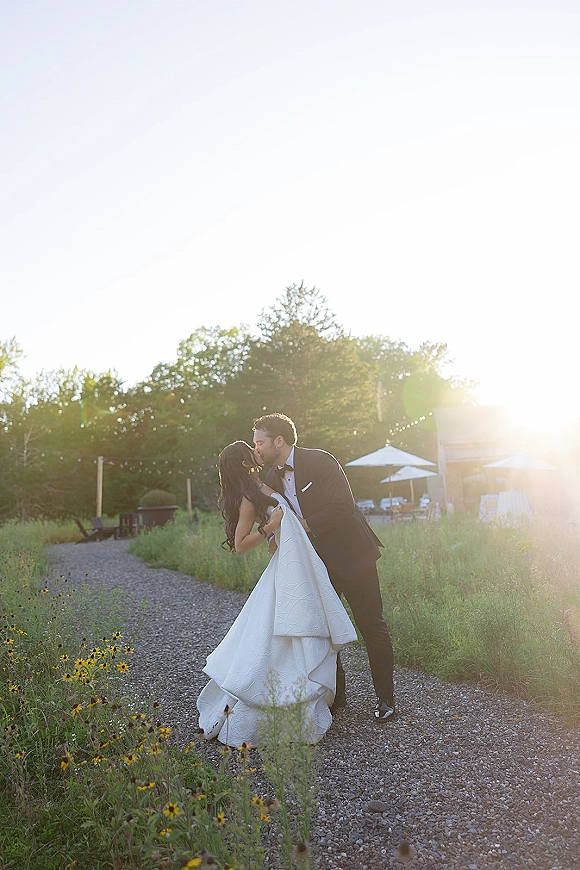 Wedding kiss as groom dips bride in a textured ball gown on a gravel path with wildflowers, string lights, and sun flare at golden hour