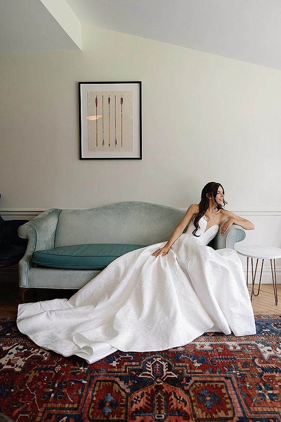 Bridal portrait of a bride in a strapless wedding dress with a long train, seated on a teal velvet sofa beside framed artwork