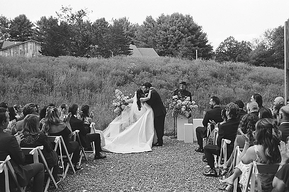 Ceremony kiss as bride in strapless gown and long veil kisses groom at outdoor altar with florals, gravel aisle, rustic buildings backdrop