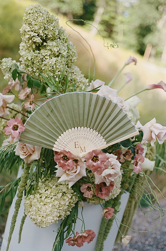 Wedding floral arrangement with hydrangea and blush roses on a ceremony chair, paired with a monogrammed hand fan in a garden setting