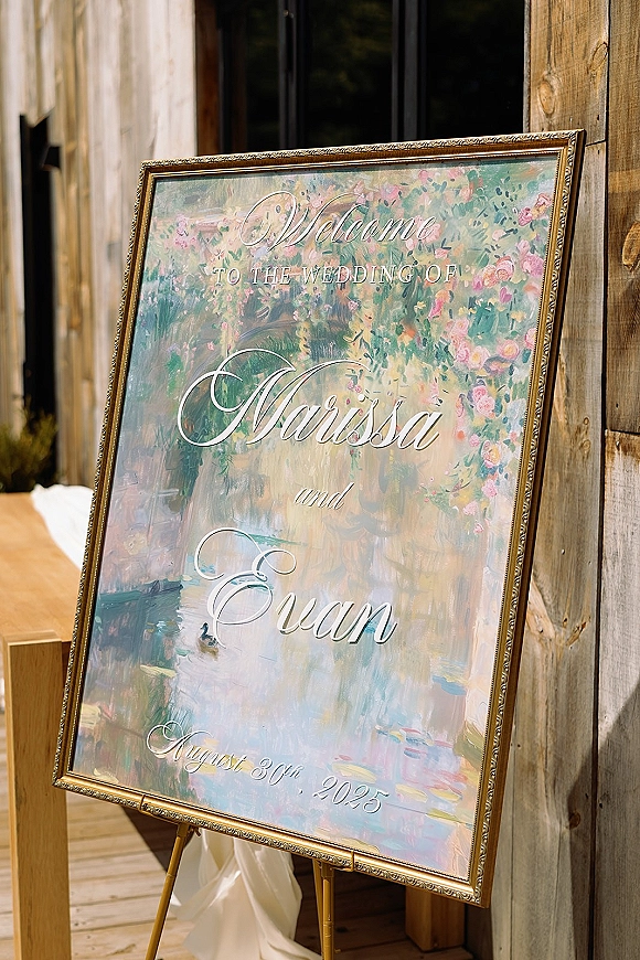 Wedding welcome sign in a gold frame with calligraphy lettering and floral artwork on an easel against a wood wall by a doorway