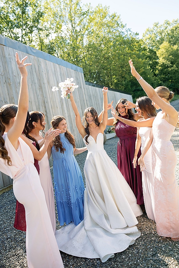 Bride with bridesmaids cheering in a bridal party celebration as she lifts a white bouquet on a gravel path by a wooden fence and trees