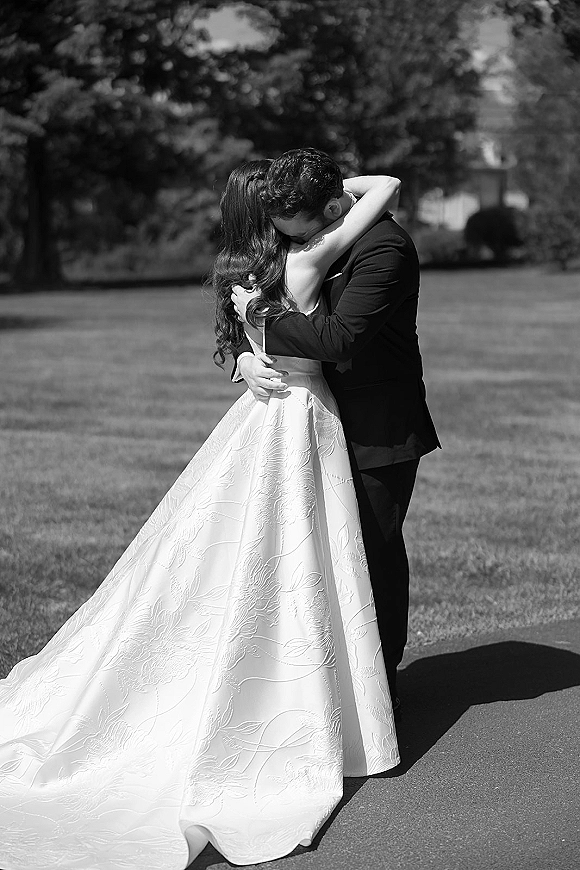Wedding kiss portrait in black and white as bride and groom kiss on a park pathway, her long wedding dress train flowing beside his tuxedo