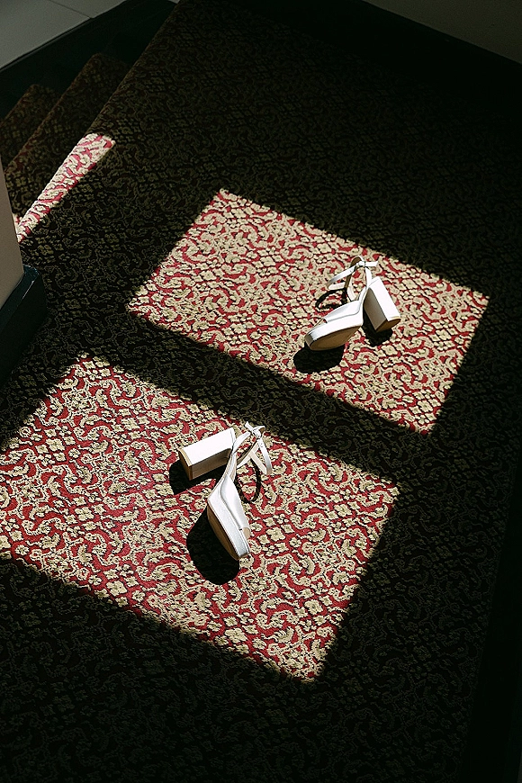 Wedding shoes in a bridal shoes flat lay: white block heel sandals with ankle straps on carpeted stairs in sunlight shadow